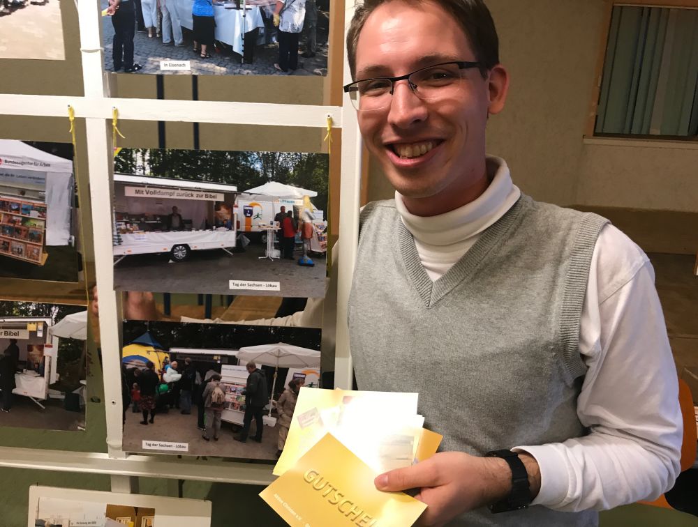 University student Samuel Naumann showing gift cards for free books beside a photo display of the book booths that his family manages in Germany. (Andrew McChesney / Adventist Mission)